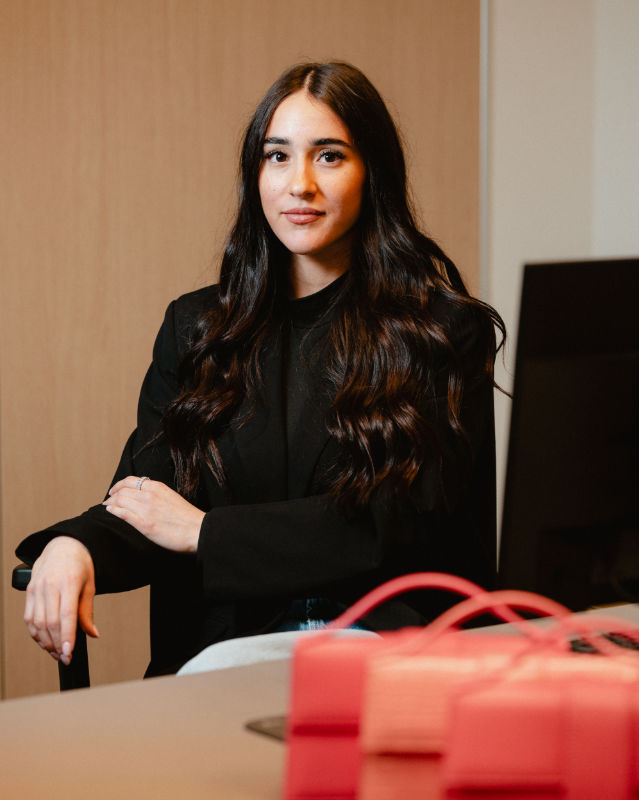 Donna seduta con le braccia incrociate accanto a una scrivania, con borse in primo piano.