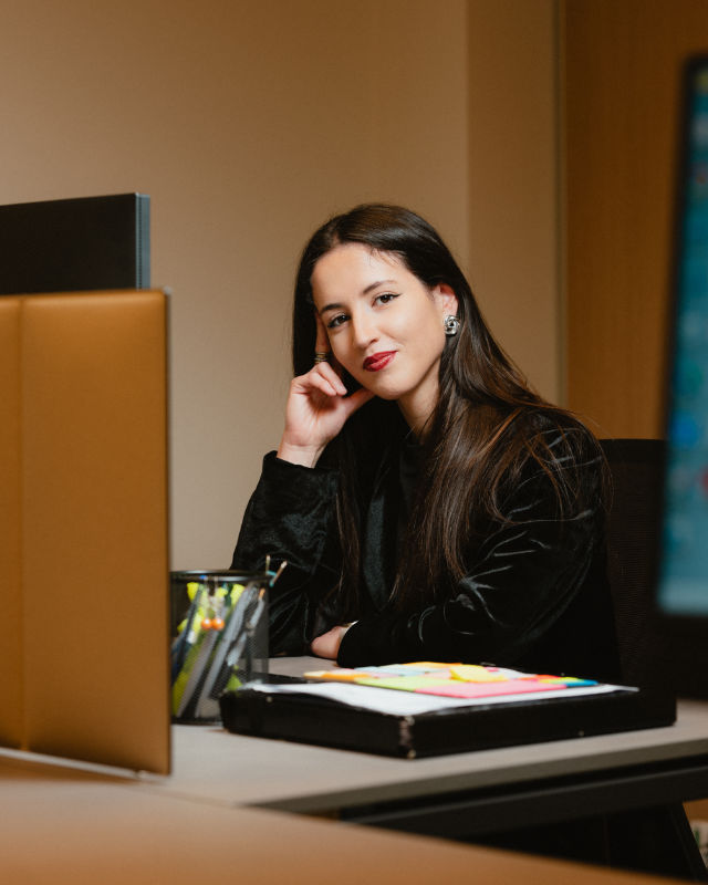 Donna seduta alla scrivania con computer, appoggia la mano al viso e guarda verso l’obiettivo.