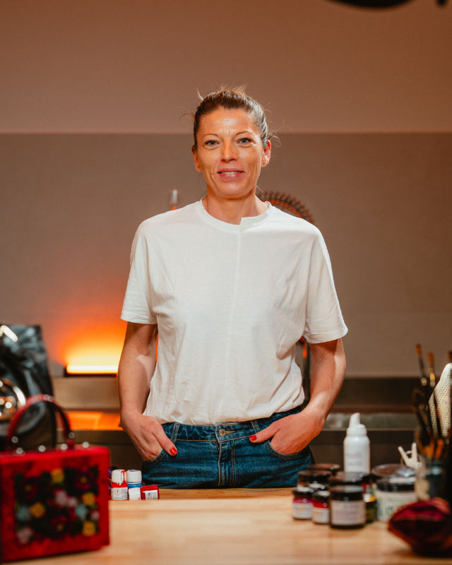 Donna in piedi dietro un banco da lavoro con prodotti e strumenti, indossa una maglietta bianca.