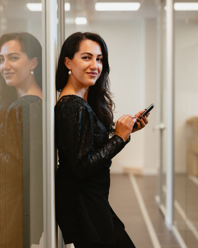 Donna sorridente appoggiata a una parete vetrata, con smartphone in mano e abito nero elegante.