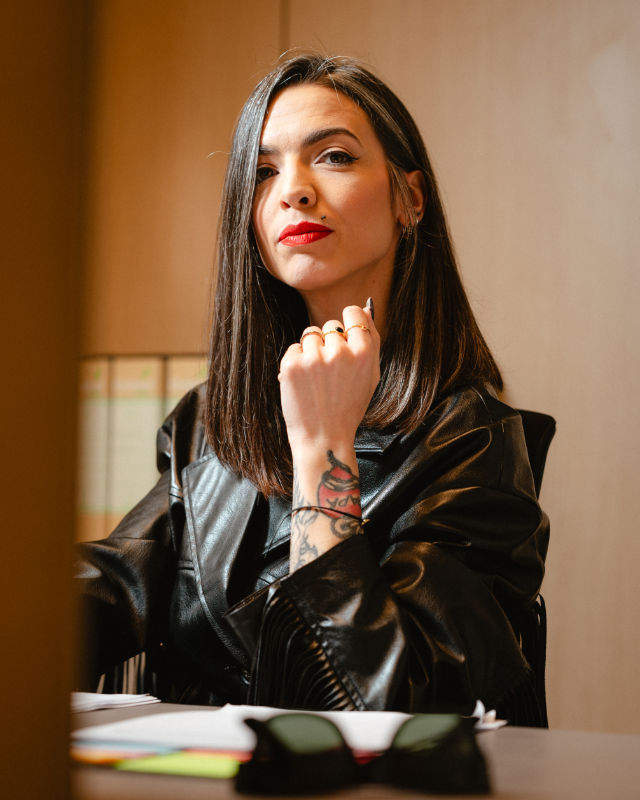 Donna con capelli lunghi scuri, in giacca di pelle nera, seduta in posa riflessiva a una scrivania.