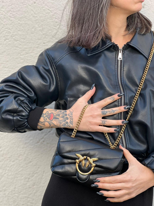 Ragazza che indossa una Borsa Pinko in pelle nera con parti metalliche dorate; munita di una tracolla scorrevole in catena con spallaccio in pelle. Indossabile a spalla oppure a tracolla. Originale, usata, di lusso, in ottime condizioni