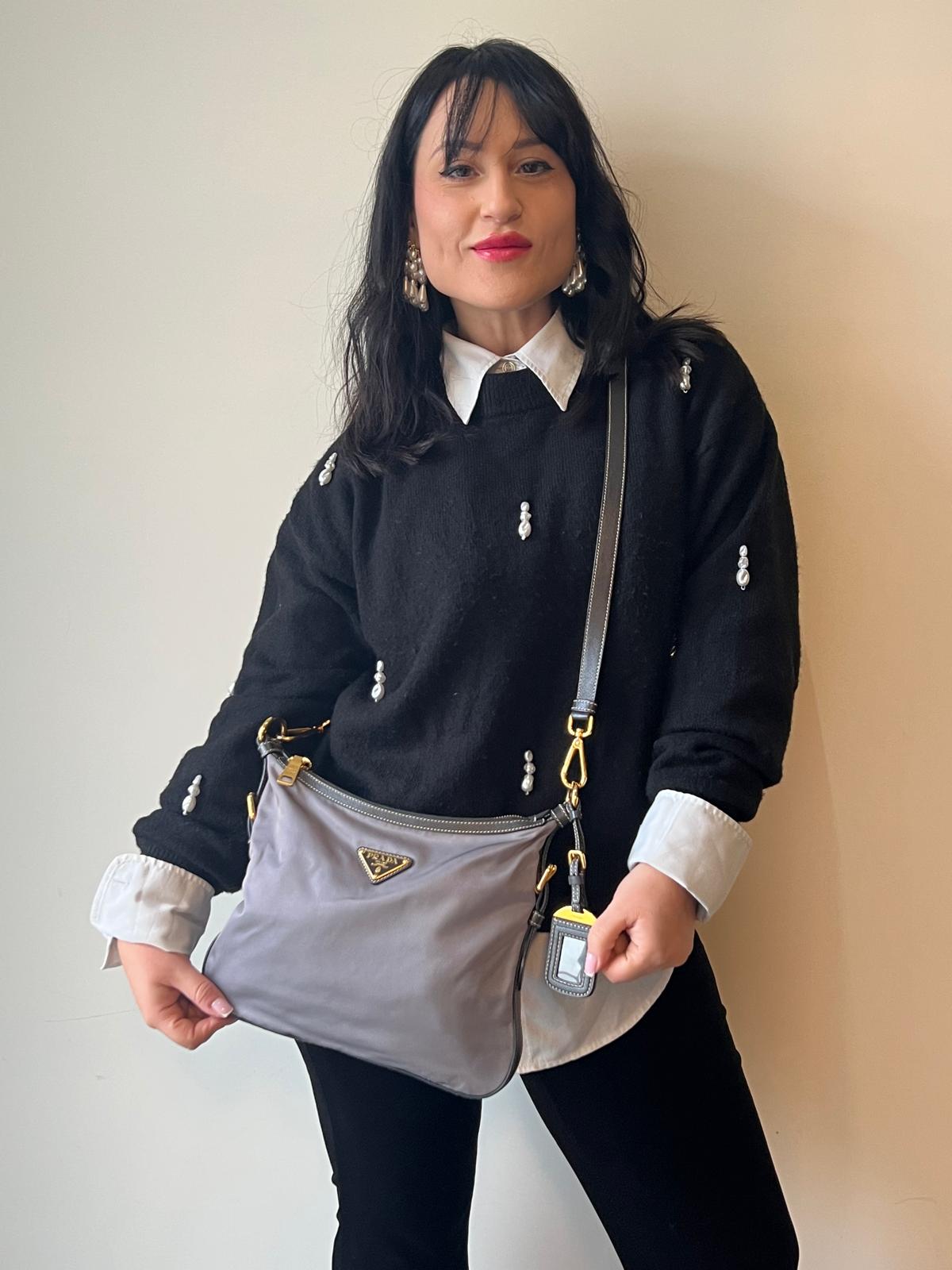 Ragazza che indossa una Borsa Prada in nylon Grigia con finiture in pelle nera e parti metalliche dorate; munita di una tracolla in tessuto regolabile e tag porta address. Originale, usata, di lusso in ottime condizioni.