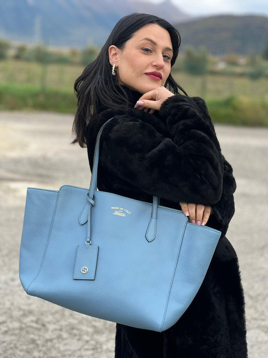Ragazza che indossa una Borsa Gucci in pelle martellata color Blu fiordaliso con incisioni dorate munita di due manici e tag porta adress. Indossabile a spalla. Completa di cartellino. Originale, usata, di lusso, in ottime condizioni.