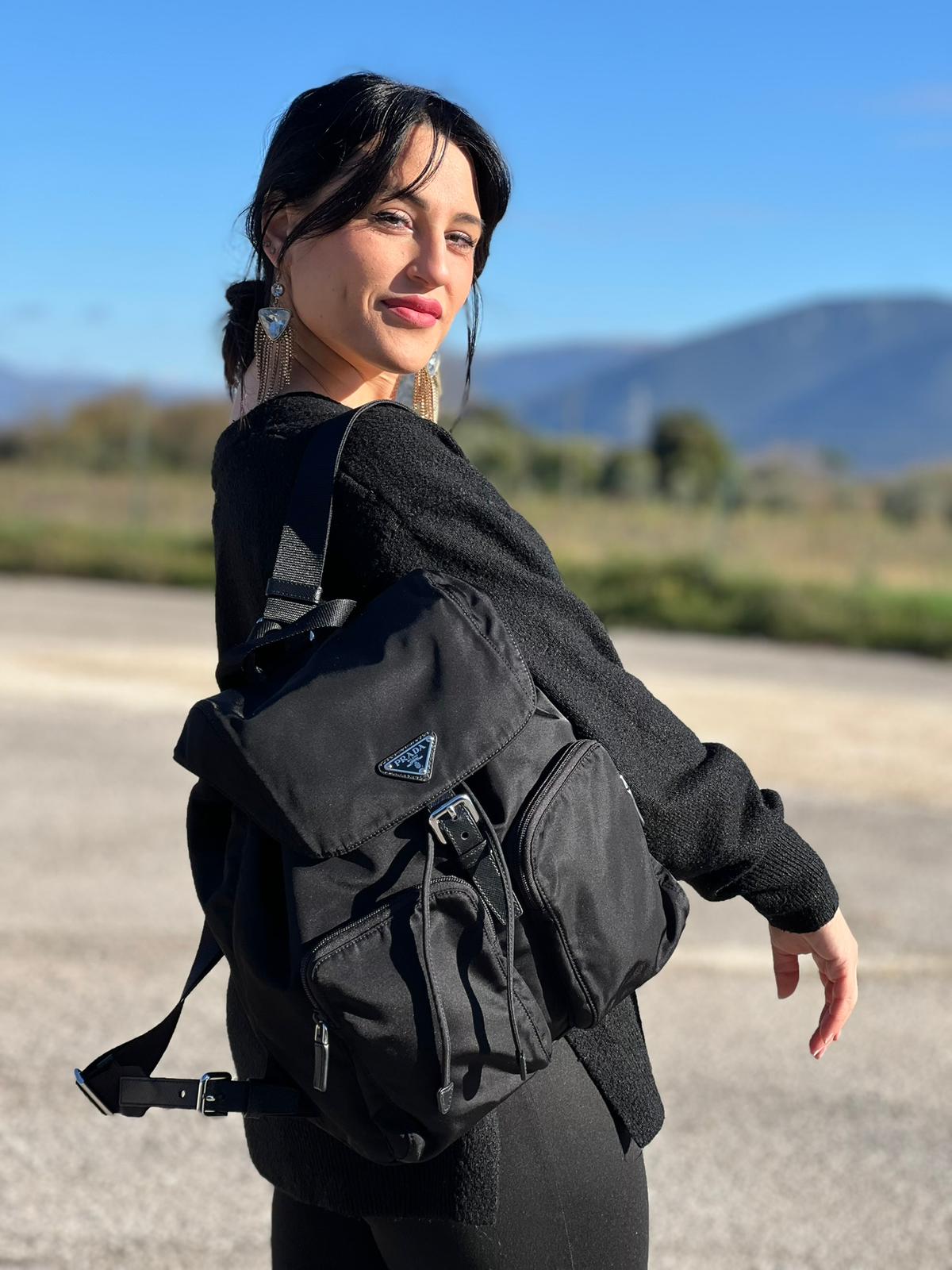 Ragazza che indossa Zaino Prada in tessuto nylon nero e finiture in pelle saffiano tono su tono. Parti metalliche argentate; munito di doppie bretelle in tessuto regolabili. Completo di corredo e card d'autenticità. Originale, usato, di lusso, in condizioni eccellenti.