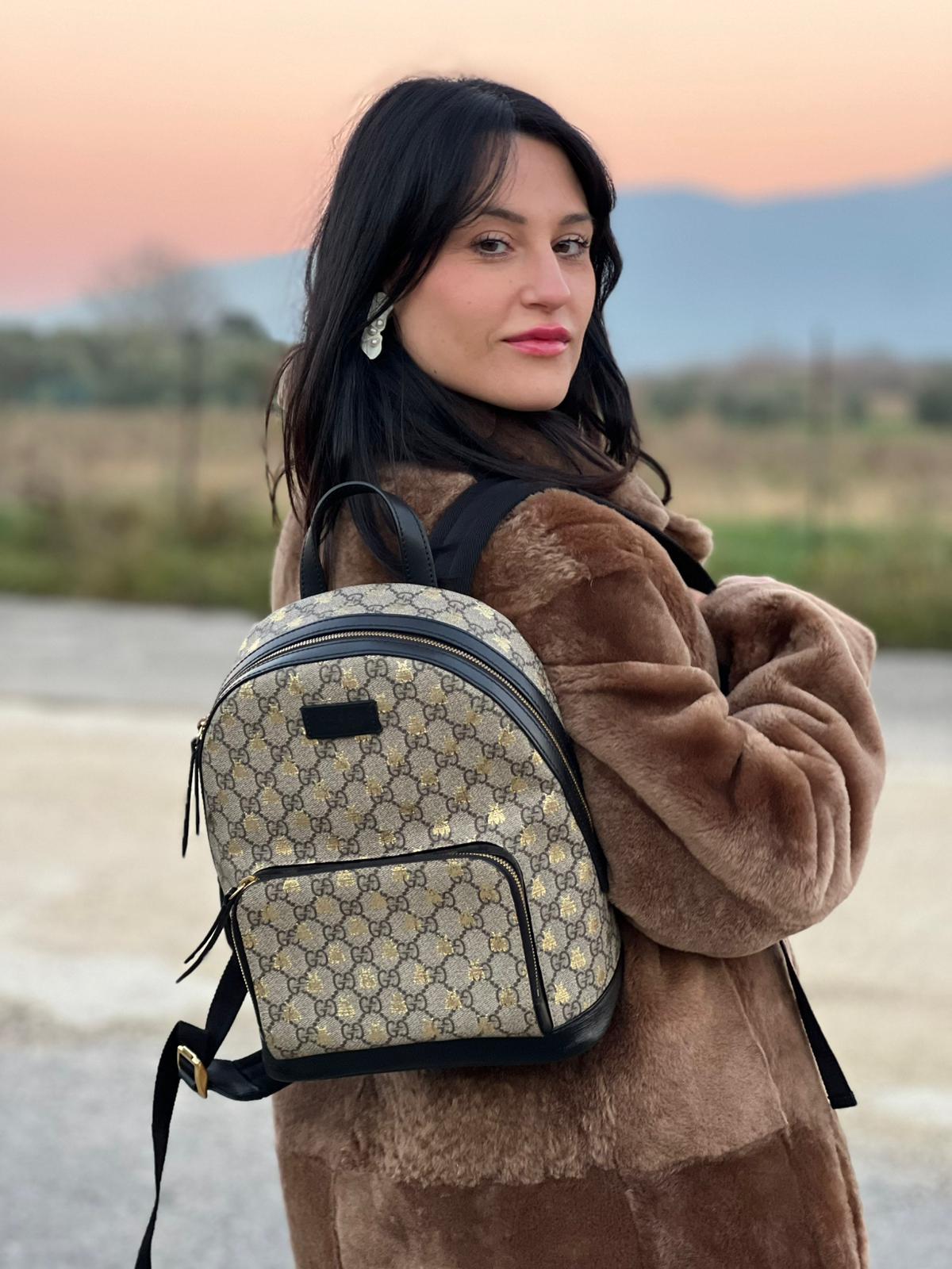 Ragazza che indossa un Zaino Gucci in canvas GG beige con finiture in pelle nere e parti metalliche dorate; presenta una stampa di api dorate su tutta la superficie, munito di un manico e bretelle in tessuto regolabili. Originale, usato, di lusso, in ottime condizioni.