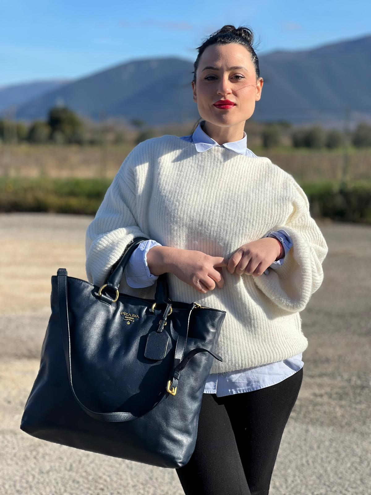 Ragazza che indossa una Borsa Prada in pelle blu notte con parti metalliche dorate; munita di un accessorio porta address, doppi manici stondati e una tracolla amovibile e regolabile. Indossabile a mano oppure a tracolla. Originale, usata, di lusso, in ottime condizioni.