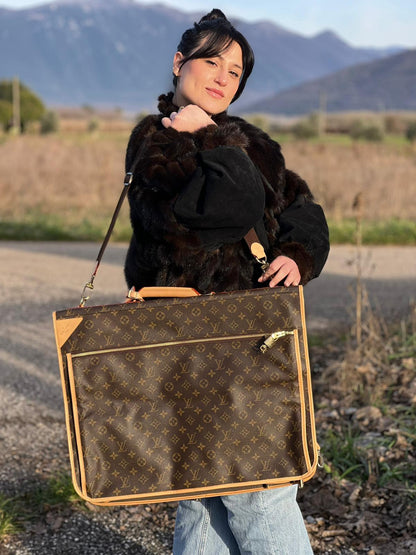 Ragazza che indossa un Borsa da viaggio porta abiti in canvas marrone monogram con finiture in vacchetta naturale e parti metalliche dorate; munita di un manico singolo, una tracolla amovibile e regolabile e 2 grucce. Completa di Lucchetti e una chiave, dustbag.