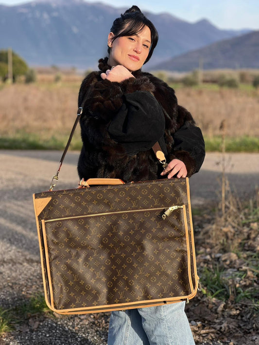 Ragazza che indossa un Borsa da viaggio porta abiti in canvas marrone monogram con finiture in vacchetta naturale e parti metalliche dorate; munita di un manico singolo, una tracolla amovibile e regolabile e 2 grucce. Completa di Lucchetti e una chiave, dustbag.