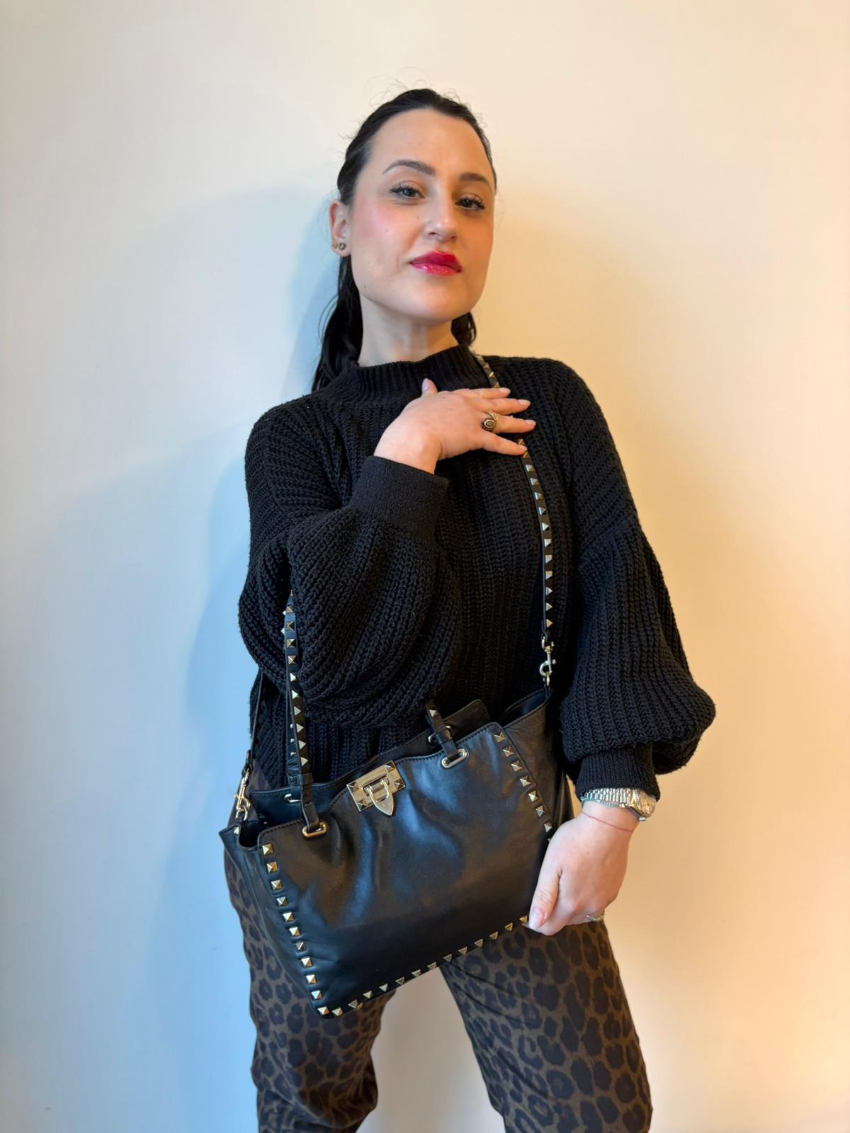 Ragazza che indossa una Borsa Valentino Garavani in pelle nera con parti metalliche dorate; impreziosita da borchie piramidali e munita di doppi manici e una tracolla amovibile. Completa di dustbag e cartellino. Originale, usata, di lusso, in ottime condizioni.