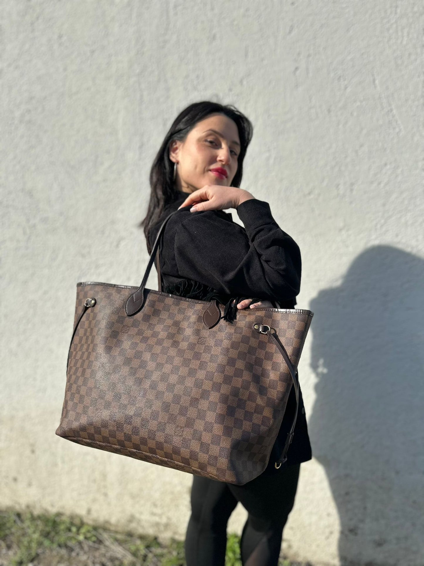 Ragazza che indossa una borsa Louis Vuitton Neverfull GM in canvas Damier Ébène con finiture in pelle marroni testa di moro e parti metalliche dorate; munita di doppi manici, indossabile a spalla.