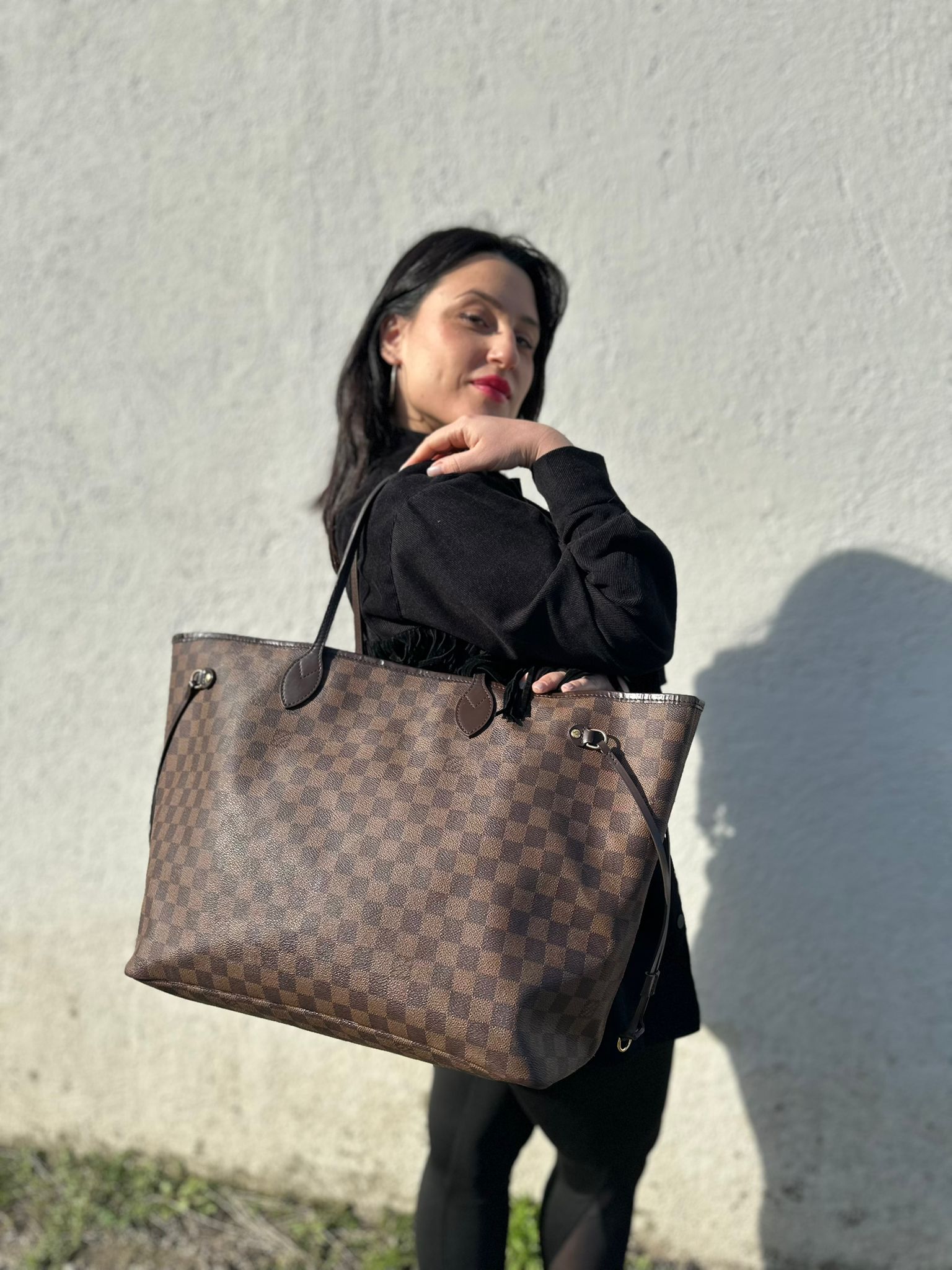 Ragazza che indossa una borsa Louis Vuitton Neverfull GM in canvas Damier Ébène con finiture in pelle marroni testa di moro e parti metalliche dorate; munita di doppi manici, indossabile a spalla.