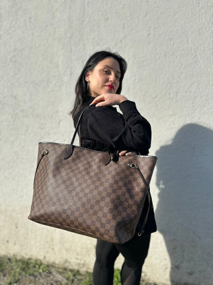 Ragazza che indossa una borsa Louis Vuitton Neverfull GM in canvas Damier Ébène con finiture in pelle marroni testa di moro e parti metalliche dorate; munita di doppi manici, indossabile a spalla.