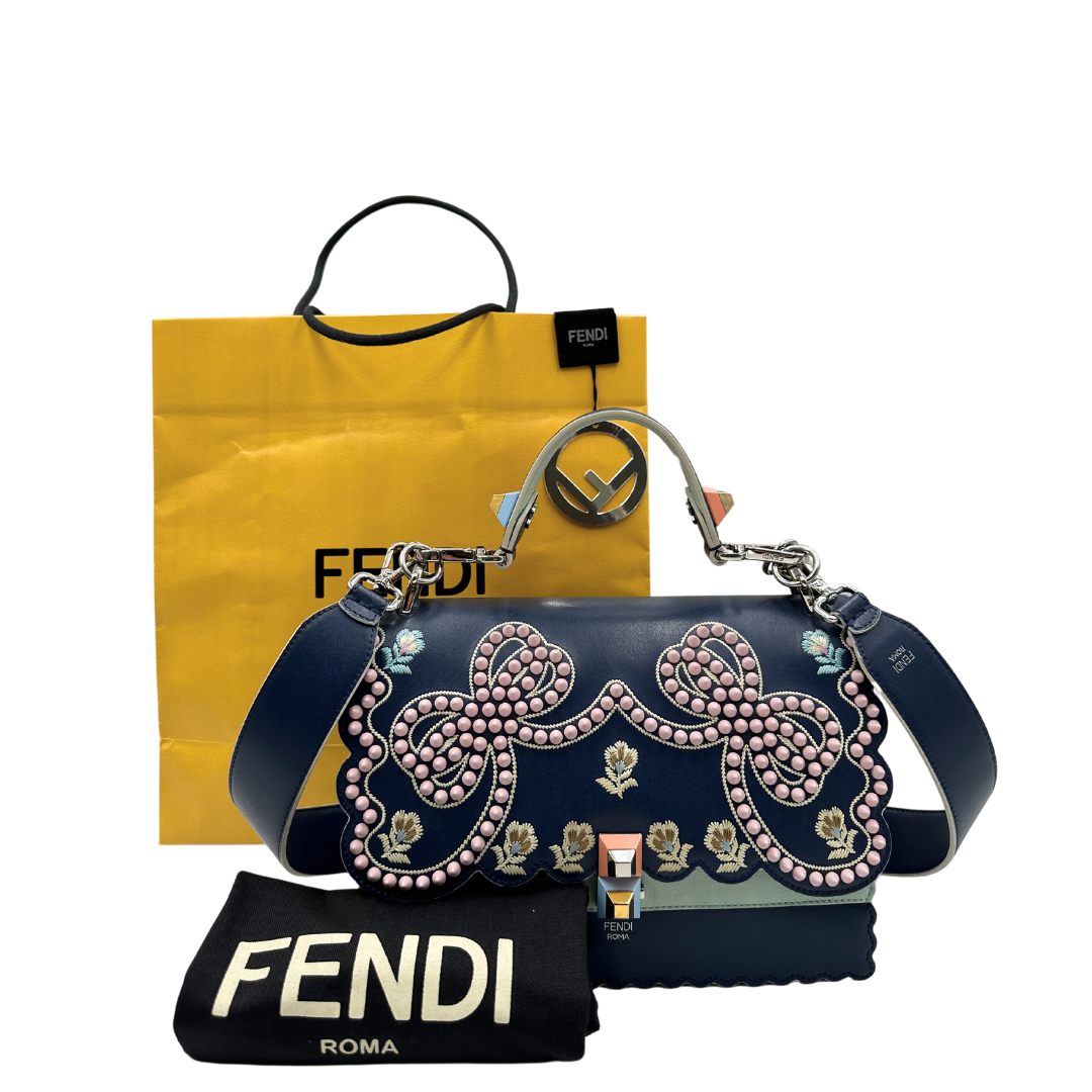 Tracolla Fendi Kan in pelle blu con dettagli floreali e borchie rosa, perfetta per aggiungere un tocco elegante e sofisticato al tuo look. Scopri il design unico e l'artigianalità Fendi.