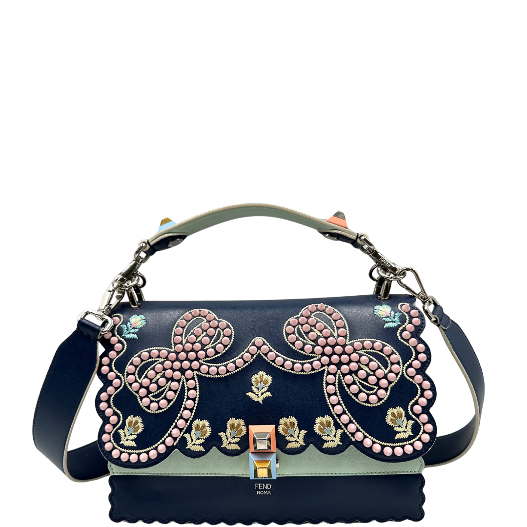 Tracolla Fendi Kan in pelle blu con dettagli floreali e borchie rosa, perfetta per aggiungere un tocco elegante e sofisticato al tuo look. Scopri il design unico e l'artigianalità Fendi.