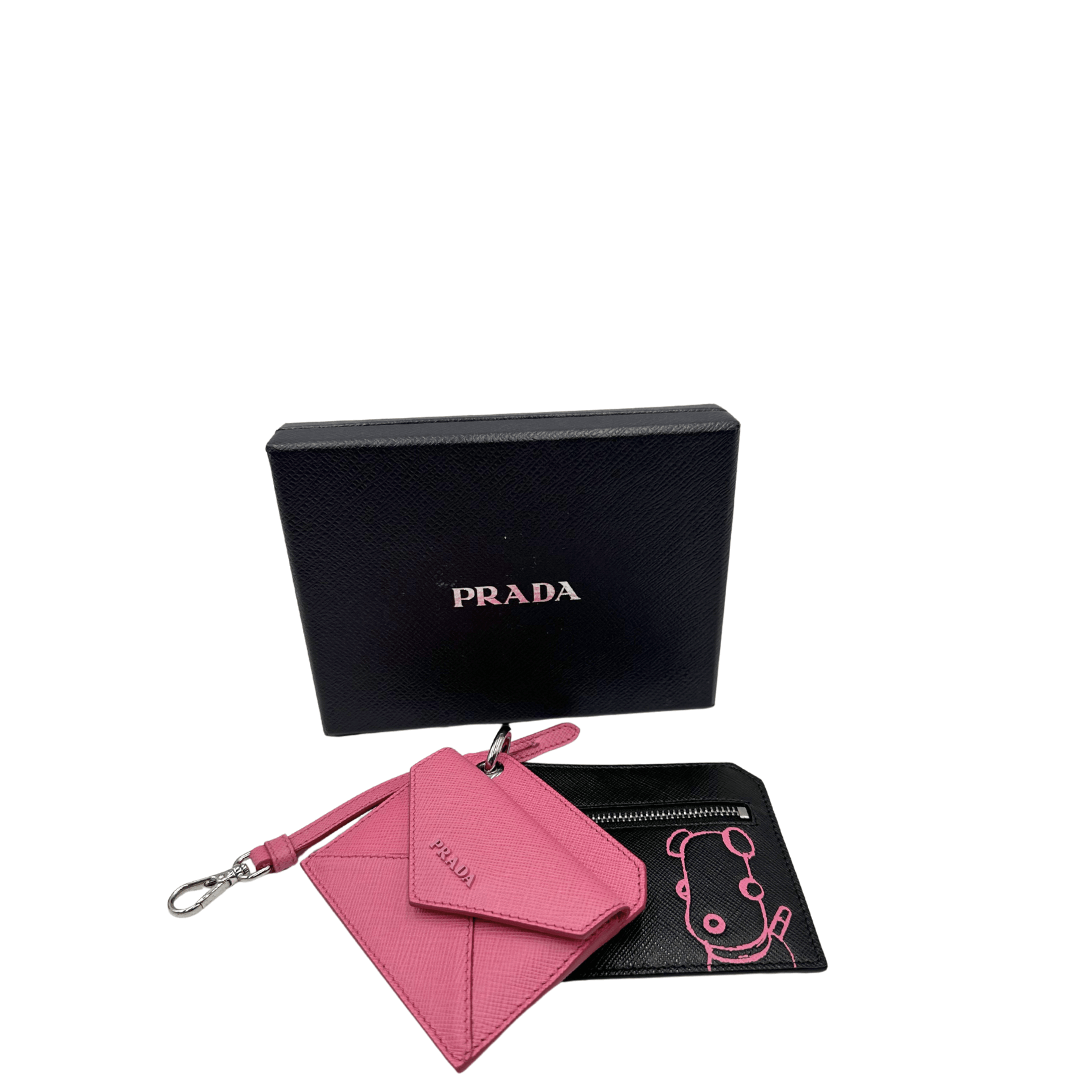 Questo set Prada include una raffinata pochette in pelle saffiano rosa con chiusura a busta e una seconda pochette nera decorata con una simpatica illustrazione. Perfetto per aggiungere un tocco di stile pratico ed elegante, rappresenta l'essenza della funzionalità e del design esclusivo Prada.