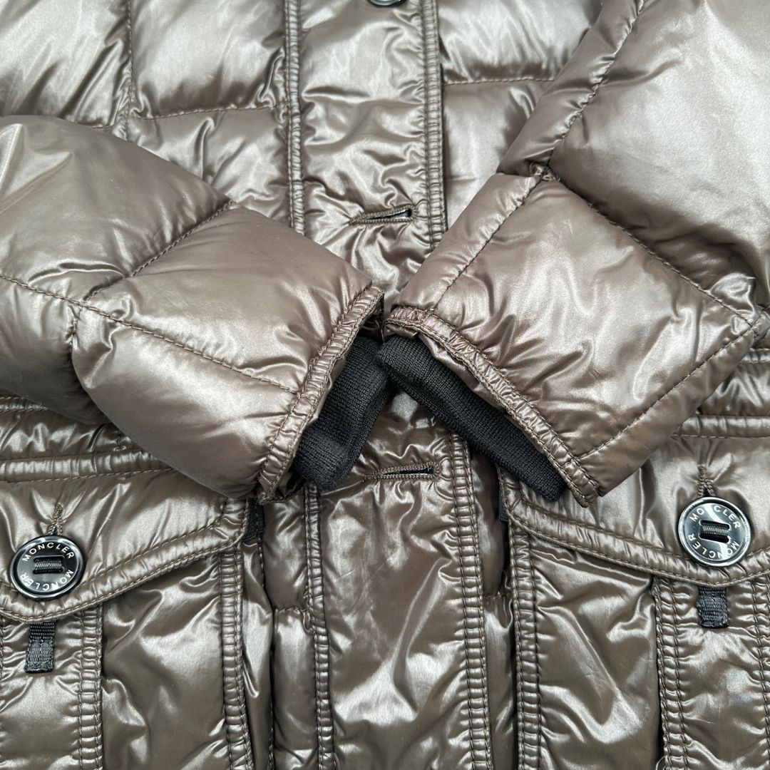 Piumino Moncler in tessuto lucido color marrone, con parti metalliche effetto renio, in 100% poliammide, caratterizzato da imbottitura trapuntata, chiusura con zip e bottoni logati, cappuccio regolabile e polsini interni a coste per una maggiore protezione dal freddo.