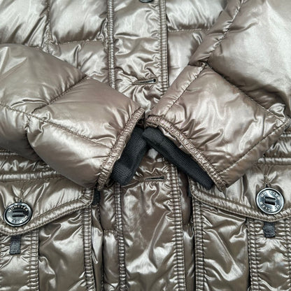 Piumino Moncler in tessuto lucido color marrone, con parti metalliche effetto renio, in 100% poliammide, caratterizzato da imbottitura trapuntata, chiusura con zip e bottoni logati, cappuccio regolabile e polsini interni a coste per una maggiore protezione dal freddo.