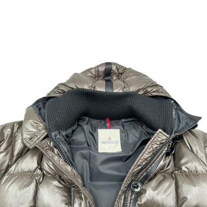 Piumino Moncler in tessuto lucido color marrone, con parti metalliche effetto renio, in 100% poliammide, caratterizzato da imbottitura trapuntata, chiusura con zip e bottoni logati, cappuccio regolabile e polsini interni a coste per una maggiore protezione dal freddo.