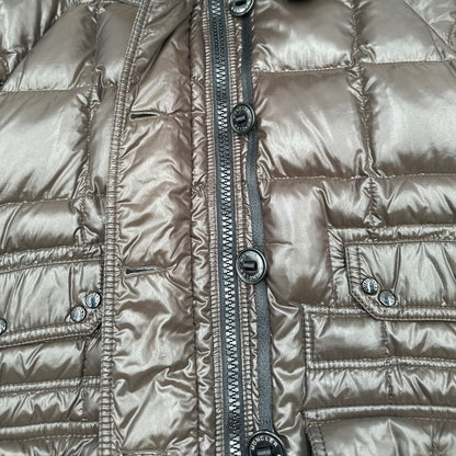 Piumino Moncler in tessuto lucido color marrone, con parti metalliche effetto renio, in 100% poliammide, caratterizzato da imbottitura trapuntata, chiusura con zip e bottoni logati, cappuccio regolabile e polsini interni a coste per una maggiore protezione dal freddo.