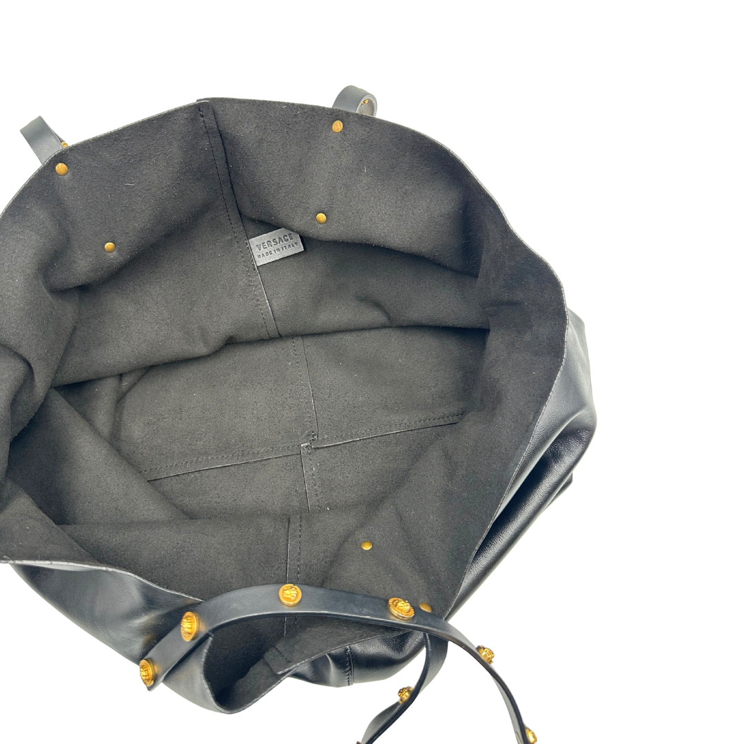 Interno di una Shopper Versace in pelle nera con parti metalliche dorate munita di due manici con borchie dorate decorative. Indossabile a spalla. N.B. Pochette mancante e punto di aggancio in pelle tagliato dalla precedente cliente. Originale, usata, di lusso, in ottime condizioni.