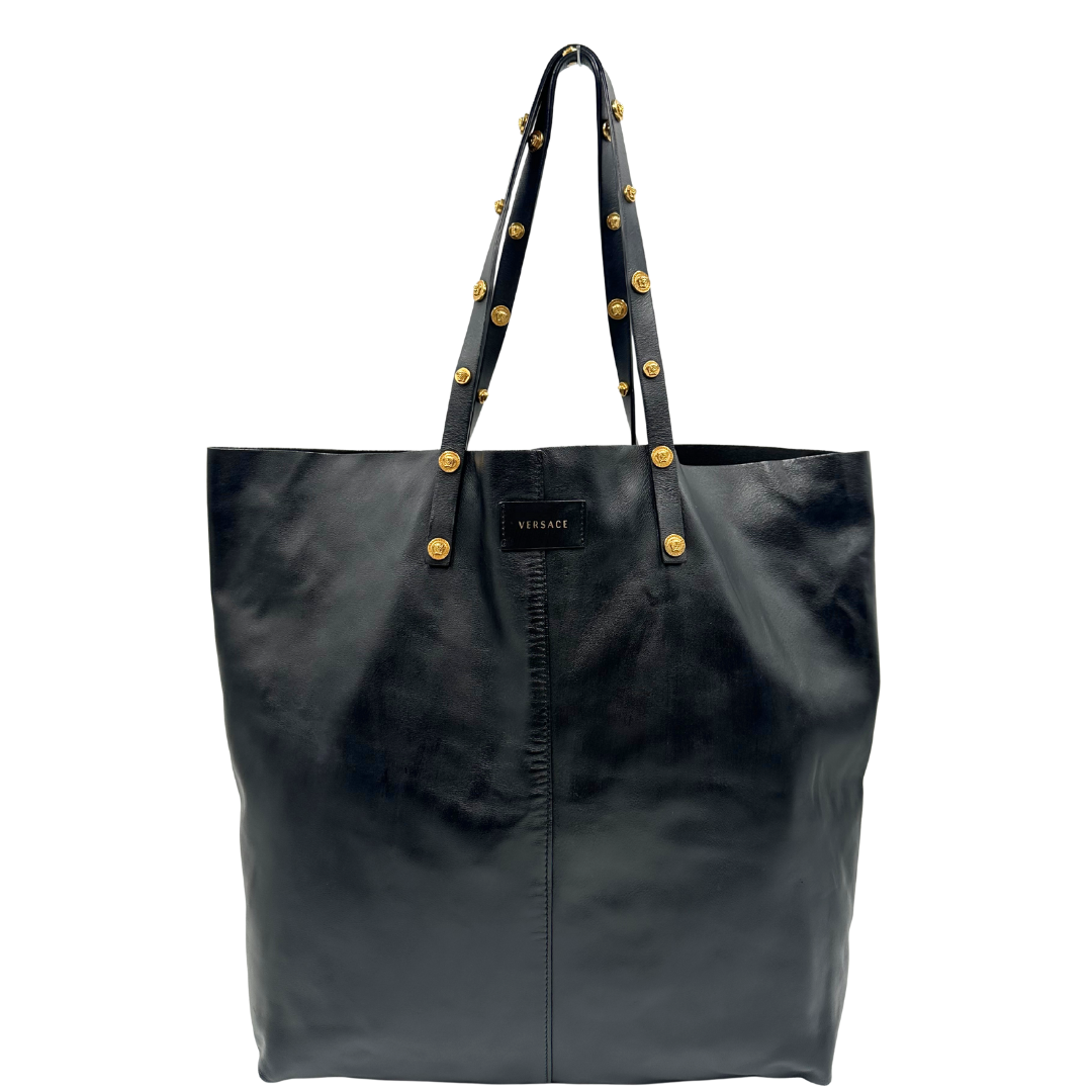 Front di una Shopper Versace in pelle nera con parti metalliche dorate munita di due manici con borchie dorate decorative. Indossabile a spalla. N.B. Pochette mancante e punto di aggancio in pelle tagliato dalla precedente cliente. Originale, usata, di lusso, in ottime condizioni.