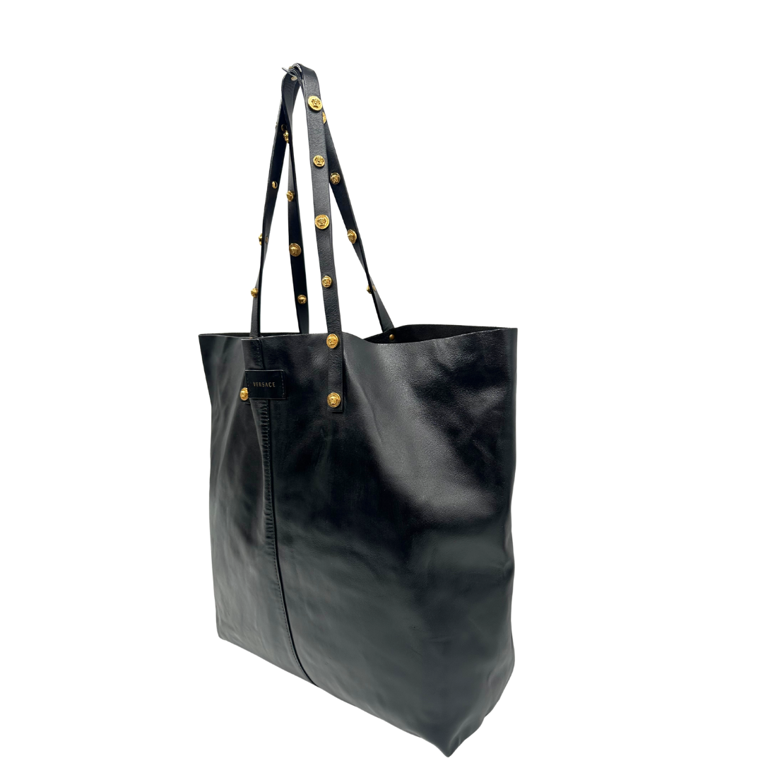 Lato sinistro di una Shopper Versace in pelle nera con parti metalliche dorate munita di due manici con borchie dorate decorative. Indossabile a spalla. N.B. Pochette mancante e punto di aggancio in pelle tagliato dalla precedente cliente. Originale, usata, di lusso, in ottime condizioni.