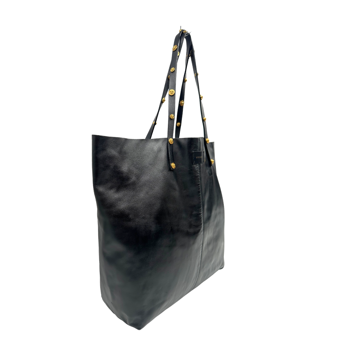 Lato destro di una Shopper Versace in pelle nera con parti metalliche dorate munita di due manici con borchie dorate decorative. Indossabile a spalla. N.B. Pochette mancante e punto di aggancio in pelle tagliato dalla precedente cliente. Originale, usata, di lusso, in ottime condizioni.