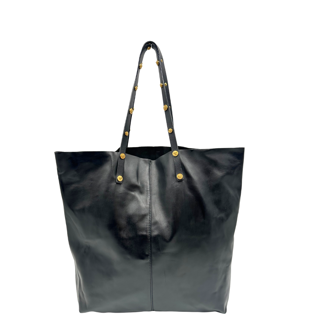 Retro di una Shopper Versace in pelle nera con parti metalliche dorate munita di due manici con borchie dorate decorative. Indossabile a spalla. N.B. Pochette mancante e punto di aggancio in pelle tagliato dalla precedente cliente. Originale, usata, di lusso, in ottime condizioni.