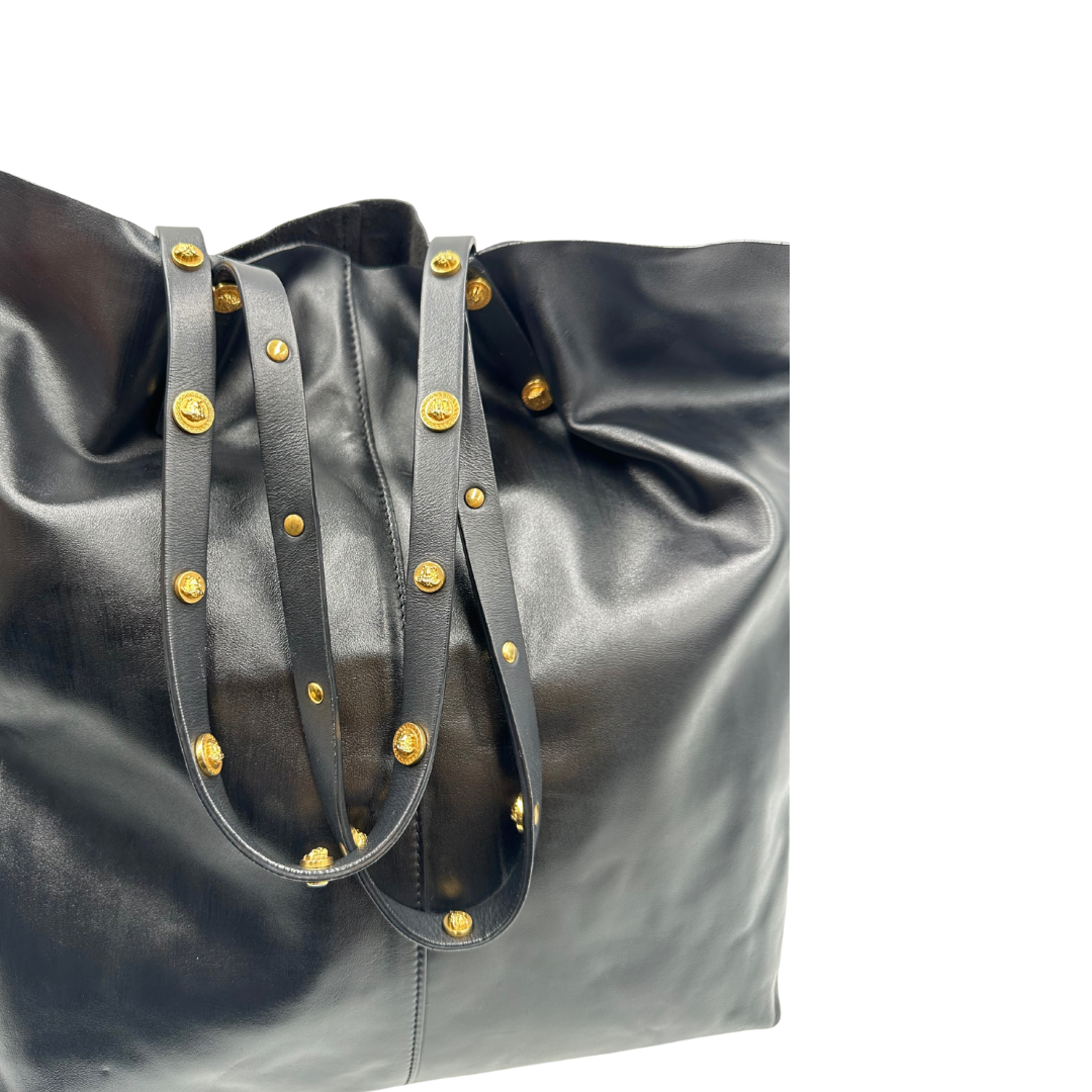 Manici di una Shopper Versace in pelle nera con parti metalliche dorate munita di due manici con borchie dorate decorative. Indossabile a spalla. N.B. Pochette mancante e punto di aggancio in pelle tagliato dalla precedente cliente. Originale, usata, di lusso, in ottime condizioni.