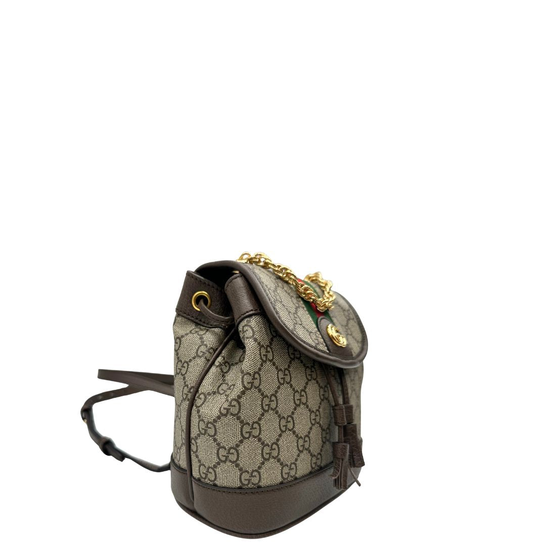 Laterale sinistro di uno zaino Gucci in canvas beige GG con finiture in pelle marroni e parti metalliche dorate. Iconica striscia Web in verde/rosso sulla patta, impreziosita da un manico in catena amovibile e cinghie in pelle regolabili. Completo di dustbag.