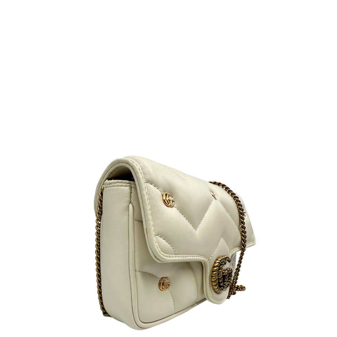 Lato destro di una Borsa Gucci GG Marmont in pelle matelassé color bianco latte, caratterizzata dalla classica trapuntatura a chevron e dagli iconici dettagli GG in metallo dorato anticato. Dotata di tracolla a catena amovibile. Presente un pouch portamonete con zip rimovibile. Completa di dustbag. Originale, usata, di lusso, in condizioni eccellenti.