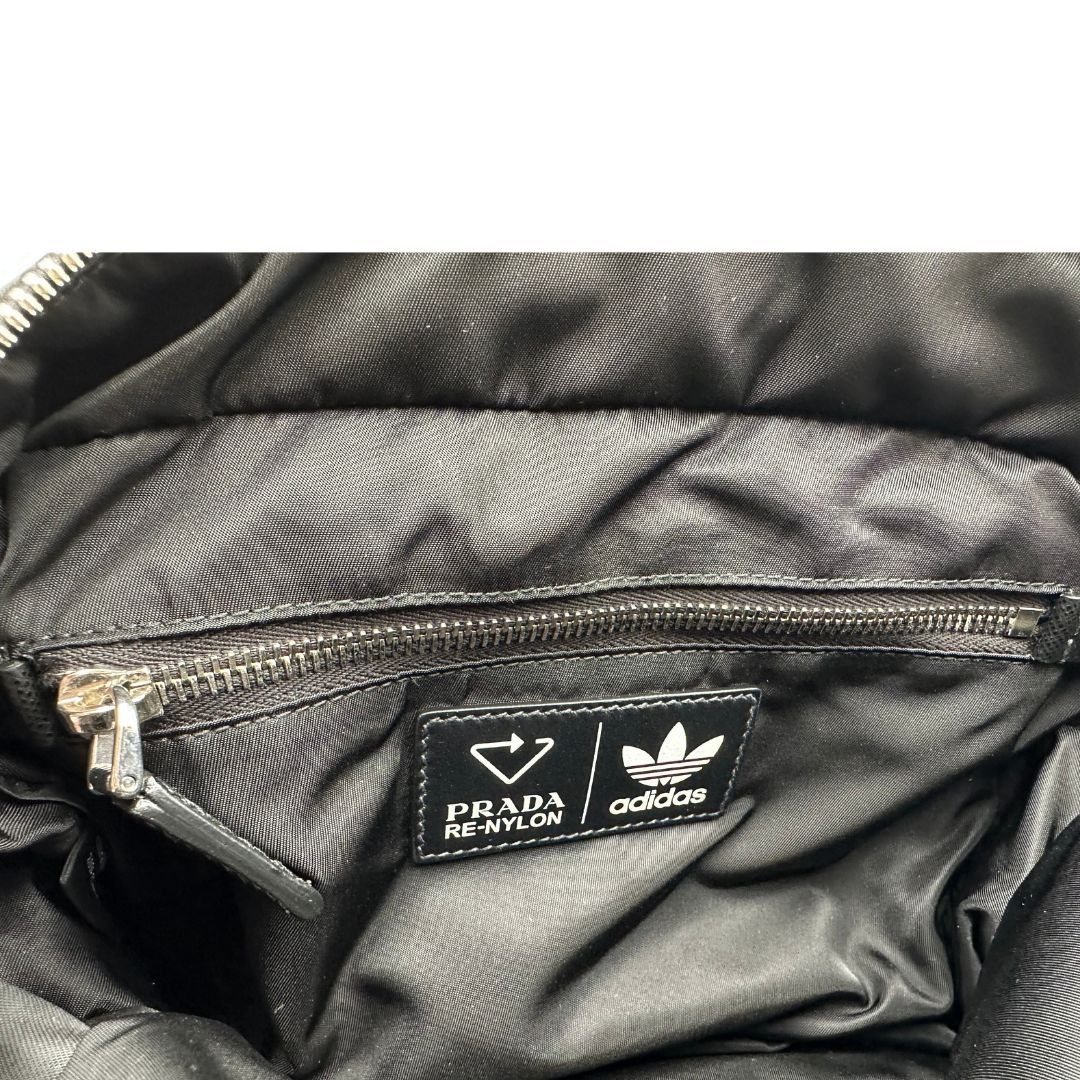 Interno di uno zaino Prada x Adidas in Re-Nylon con chiusura a zip. Struttura con tasca frontale a zip e tasca superiore integrata negli spallacci. Spallacci regolabili con imbottitura e pannello posteriore in mesh. Dettagli con logo triangolo Prada sul lato superiore e branding Prada Re-Nylon e Adidas sul fianco.