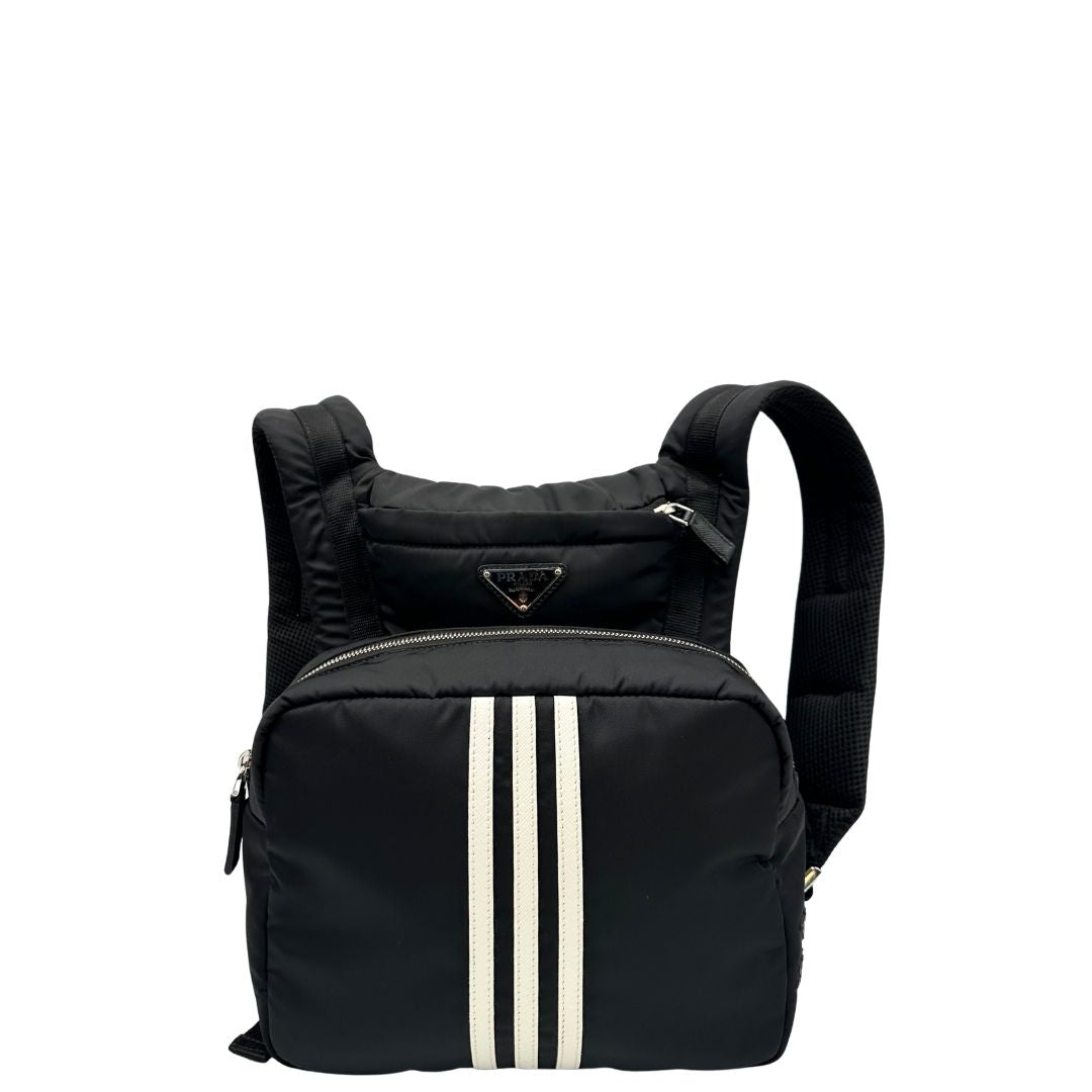 Front di uno zaino Prada x Adidas in Re-Nylon con chiusura a zip. Struttura con tasca frontale a zip e tasca superiore integrata negli spallacci. Spallacci regolabili con imbottitura e pannello posteriore in mesh. Dettagli con logo triangolo Prada sul lato superiore e branding Prada Re-Nylon e Adidas sul fianco.