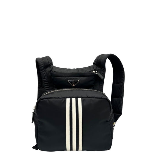 Front di uno zaino Prada x Adidas in Re-Nylon con chiusura a zip. Struttura con tasca frontale a zip e tasca superiore integrata negli spallacci. Spallacci regolabili con imbottitura e pannello posteriore in mesh. Dettagli con logo triangolo Prada sul lato superiore e branding Prada Re-Nylon e Adidas sul fianco.