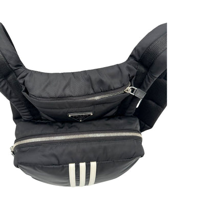 Vista superiore di uno zaino Prada x Adidas in Re-Nylon con chiusura a zip. Struttura con tasca frontale a zip e tasca superiore integrata negli spallacci. Spallacci regolabili con imbottitura e pannello posteriore in mesh. Dettagli con logo triangolo Prada sul lato superiore e branding Prada Re-Nylon e Adidas sul fianco.