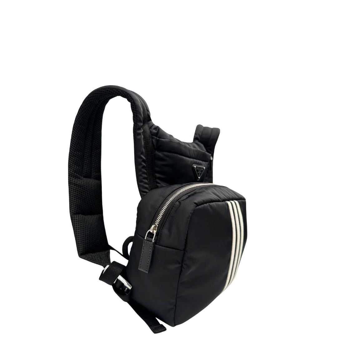 Laterale sinistro di uno zaino Prada x Adidas in Re-Nylon con chiusura a zip. Struttura con tasca frontale a zip e tasca superiore integrata negli spallacci. Spallacci regolabili con imbottitura e pannello posteriore in mesh. Dettagli con logo triangolo Prada sul lato superiore e branding Prada Re-Nylon e Adidas sul fianco.