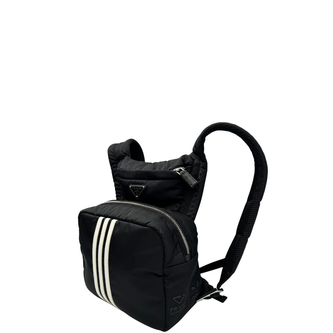 Laterale destro di uno zaino Prada x Adidas in Re-Nylon con chiusura a zip. Struttura con tasca frontale a zip e tasca superiore integrata negli spallacci. Spallacci regolabili con imbottitura e pannello posteriore in mesh. Dettagli con logo triangolo Prada sul lato superiore e branding Prada Re-Nylon e Adidas sul fianco.