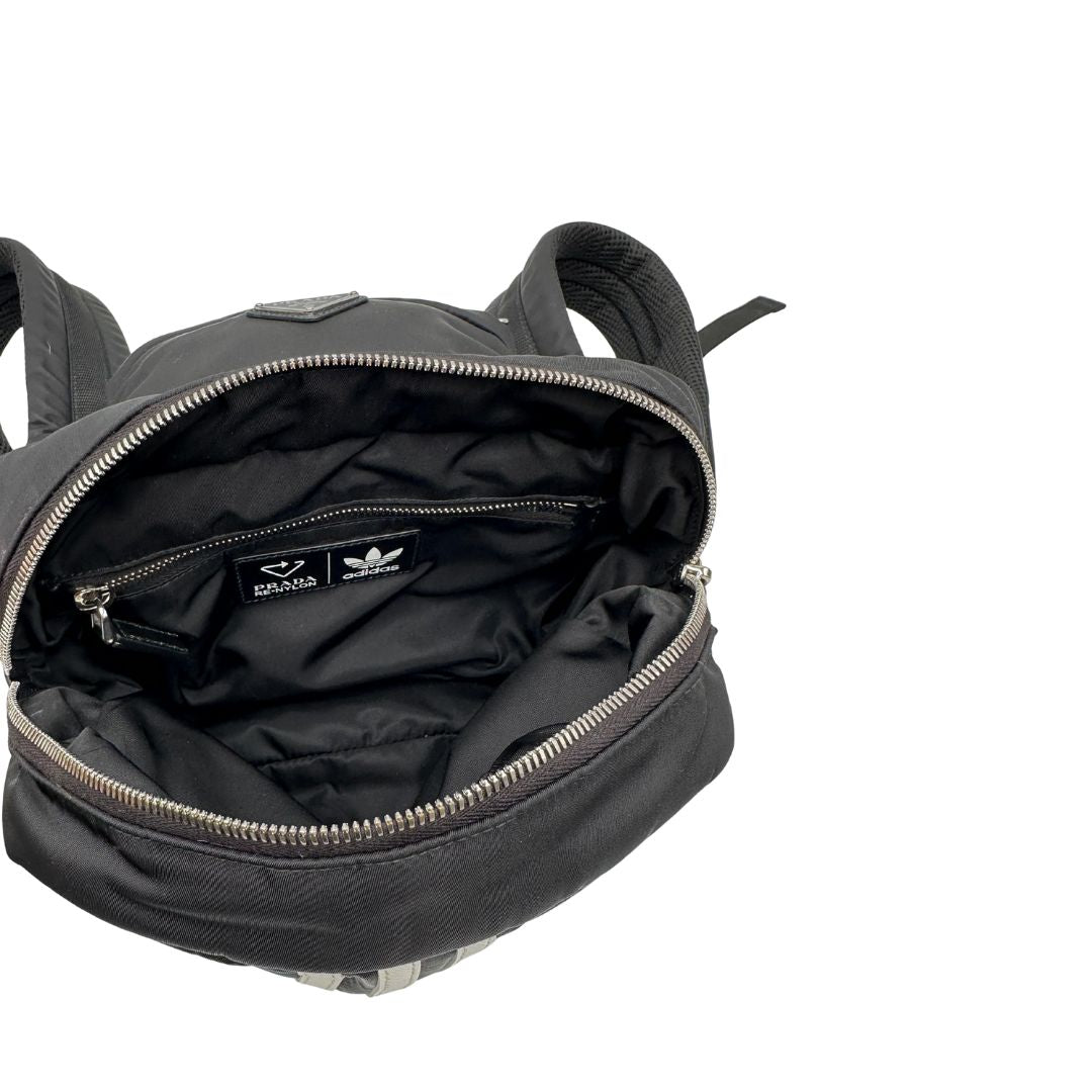 Interno di uno zaino Prada x Adidas in Re-Nylon con chiusura a zip. Struttura con tasca frontale a zip e tasca superiore integrata negli spallacci. Spallacci regolabili con imbottitura e pannello posteriore in mesh. Dettagli con logo triangolo Prada sul lato superiore e branding Prada Re-Nylon e Adidas sul fianco.