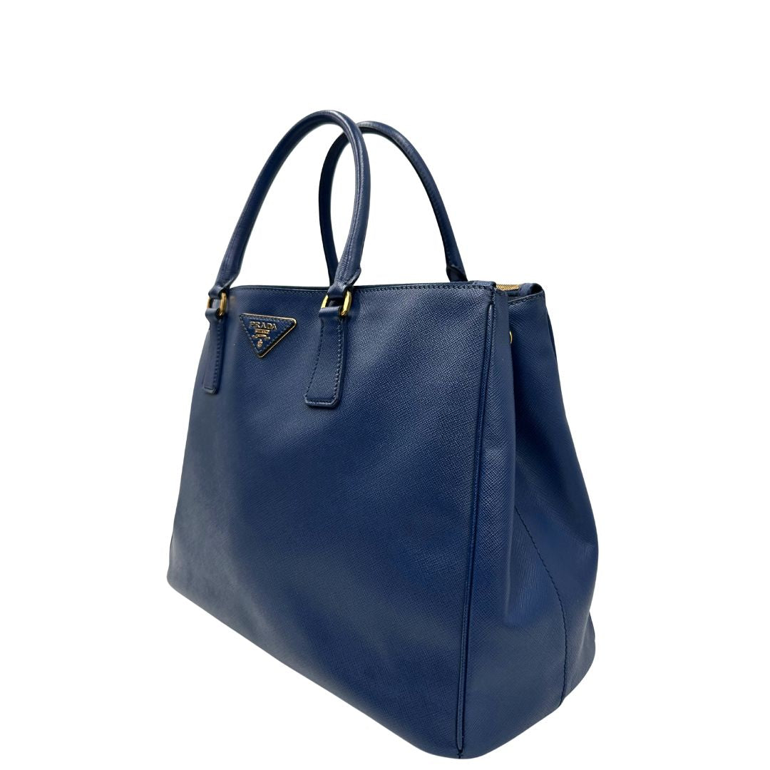 lato Borsa Prada in pelle saffiano blu con parti metalliche dorate; munita di doppi manici stondati e impreziosita da una clochette portachiavi, di lusso, originale, ottime condizioni. 