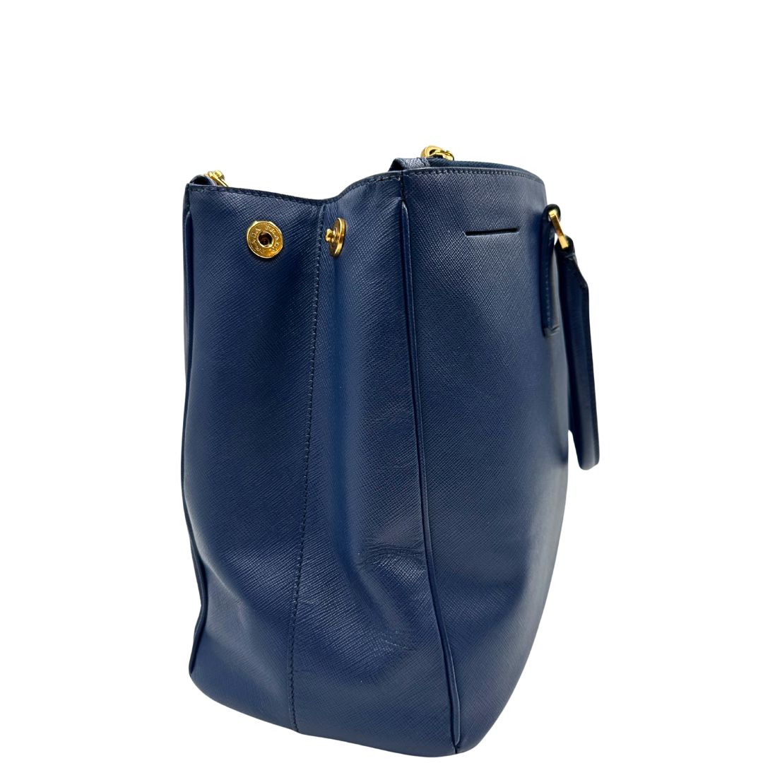 lato Borsa Prada in pelle saffiano blu con parti metalliche dorate; munita di doppi manici stondati e impreziosita da una clochette portachiavi, di lusso, originale, ottime condizioni.