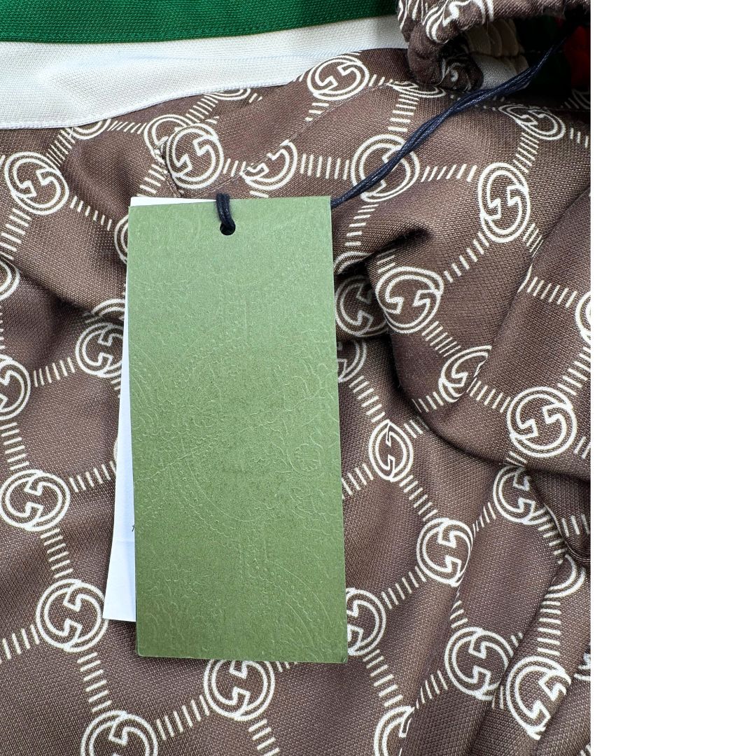 Pantalone tuta Gucci elasticizzato modello Interlocking G in 55% poliestere e 45% cotone, marrone e avorio con motivo GG all-over e bande laterali con bottoni automatici che si aprono fino ai fianchi, con strisce Web verde/rosse.