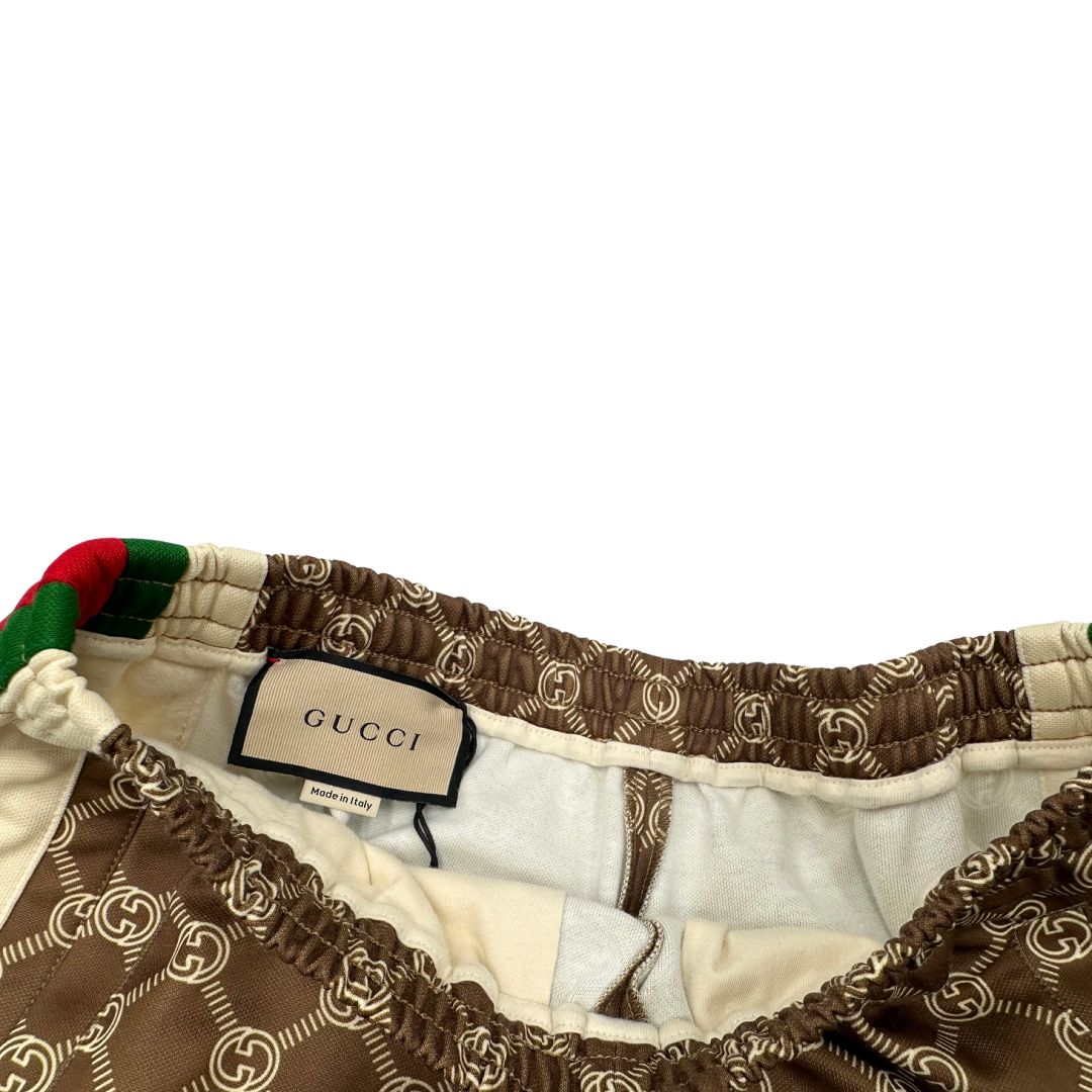 Pantalone tuta Gucci elasticizzato modello Interlocking G in 55% poliestere e 45% cotone, marrone e avorio con motivo GG all-over e bande laterali con bottoni automatici che si aprono fino ai fianchi, con strisce Web verde/rosse.