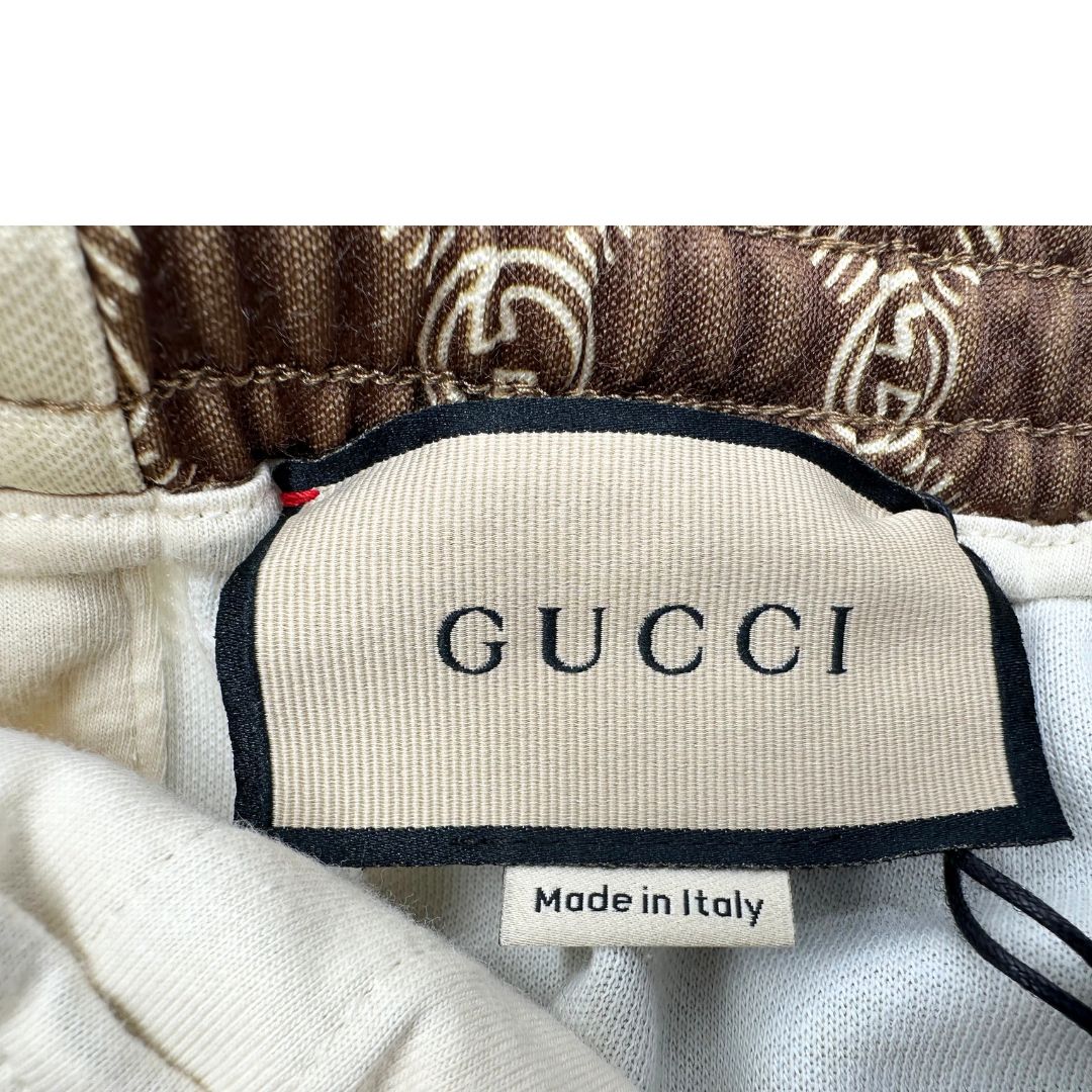 Pantalone tuta Gucci elasticizzato modello Interlocking G in 55% poliestere e 45% cotone, marrone e avorio con motivo GG all-over e bande laterali con bottoni automatici che si aprono fino ai fianchi, con strisce Web verde/rosse.