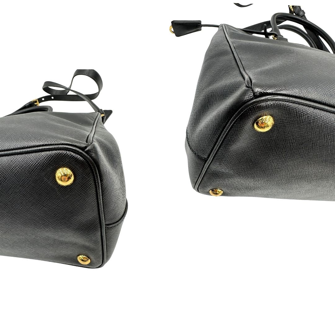 Angoli di una borsa Prada in pelle saffiano nera con parti metalliche dorate, munita di doppi manici stondati, tracolla amovibile e regolabile ed una clochette portachiavi. Completa di dustbag