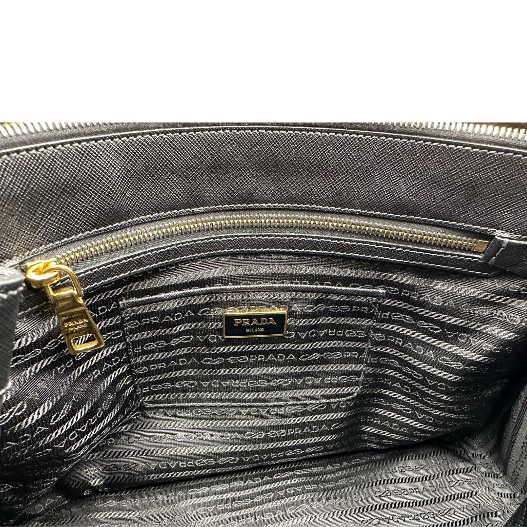 Interno di una borsa Prada in pelle saffiano nera con parti metalliche dorate, munita di doppi manici stondati, tracolla amovibile e regolabile ed una clochette portachiavi. Completa di dustbag