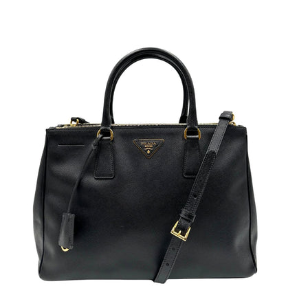 Front di una borsa Prada in pelle saffiano nera con parti metalliche dorate, munita di doppi manici stondati, tracolla amovibile e regolabile ed una clochette portachiavi. Completa di dustbag