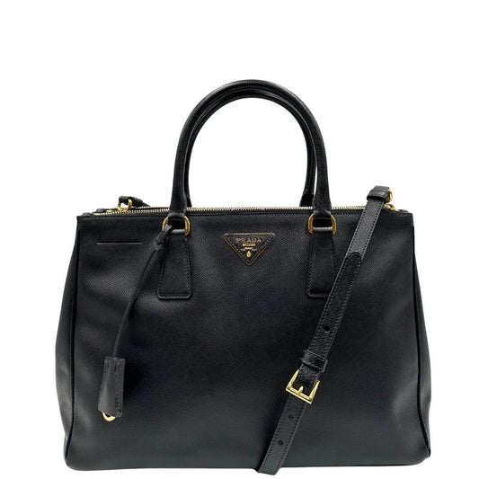 Front di una borsa Prada in pelle saffiano nera con parti metalliche dorate, munita di doppi manici stondati, tracolla amovibile e regolabile ed una clochette portachiavi. Completa di dustbag