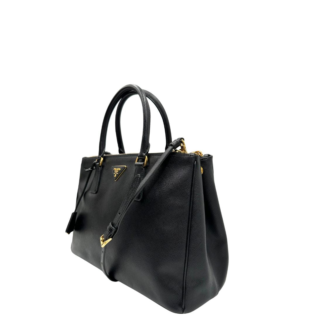 Laterale destro di una borsa Prada in pelle saffiano nera con parti metalliche dorate, munita di doppi manici stondati, tracolla amovibile e regolabile ed una clochette portachiavi. Completa di dustbag