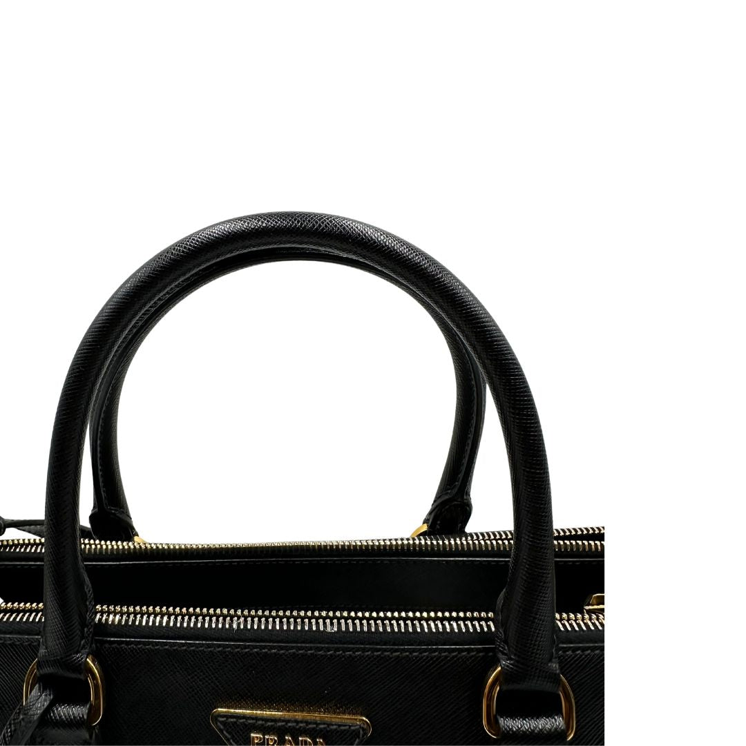 Manici di una borsa Prada in pelle saffiano nera con parti metalliche dorate, munita di doppi manici stondati, tracolla amovibile e regolabile ed una clochette portachiavi. Completa di dustbag